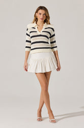 A woman is wearing a knitted sweater with horizontal black and cream stripes, featuring a fold-over collar and three-quarter length sleeves. She pairs it with a white, ruffled pleated skirt and white open-toe high-heeled sandals.
